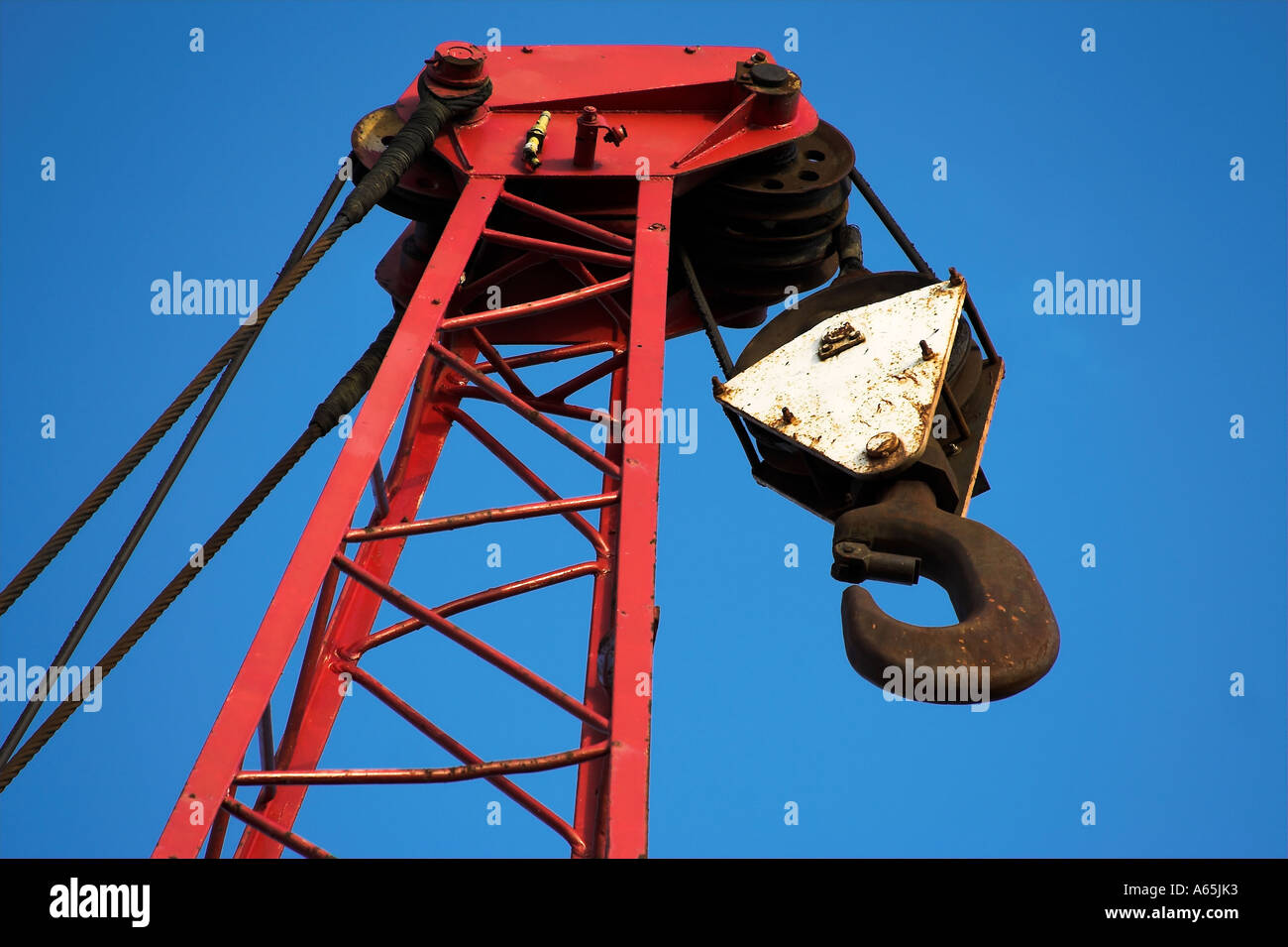 A close up view of the top of a crane with hook, cable and hoist wheels against a clear blue sky Stock Photo