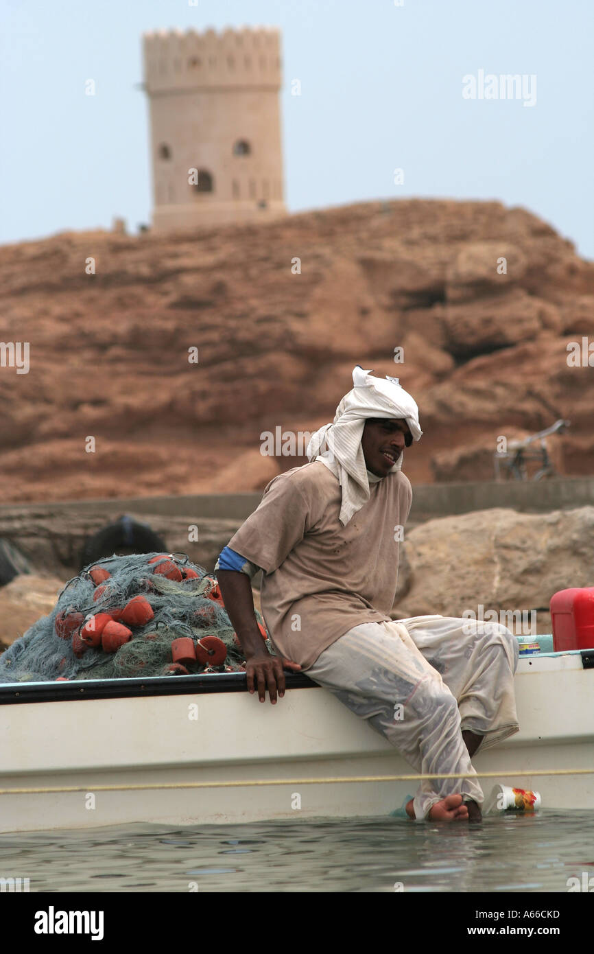 Indian fisherman on a boat in front of one of the Ayajh's defensive watchtowers on hills in Sur, Oman Stock Photo