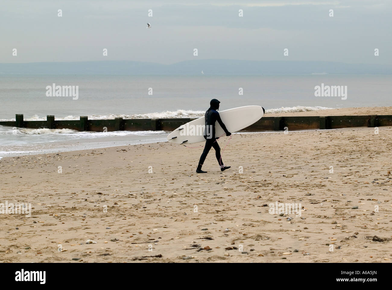 Surfer carrying his surfboard across the beach dressed in his winter wetsuit at the end of a dull winter day Stock Photo
