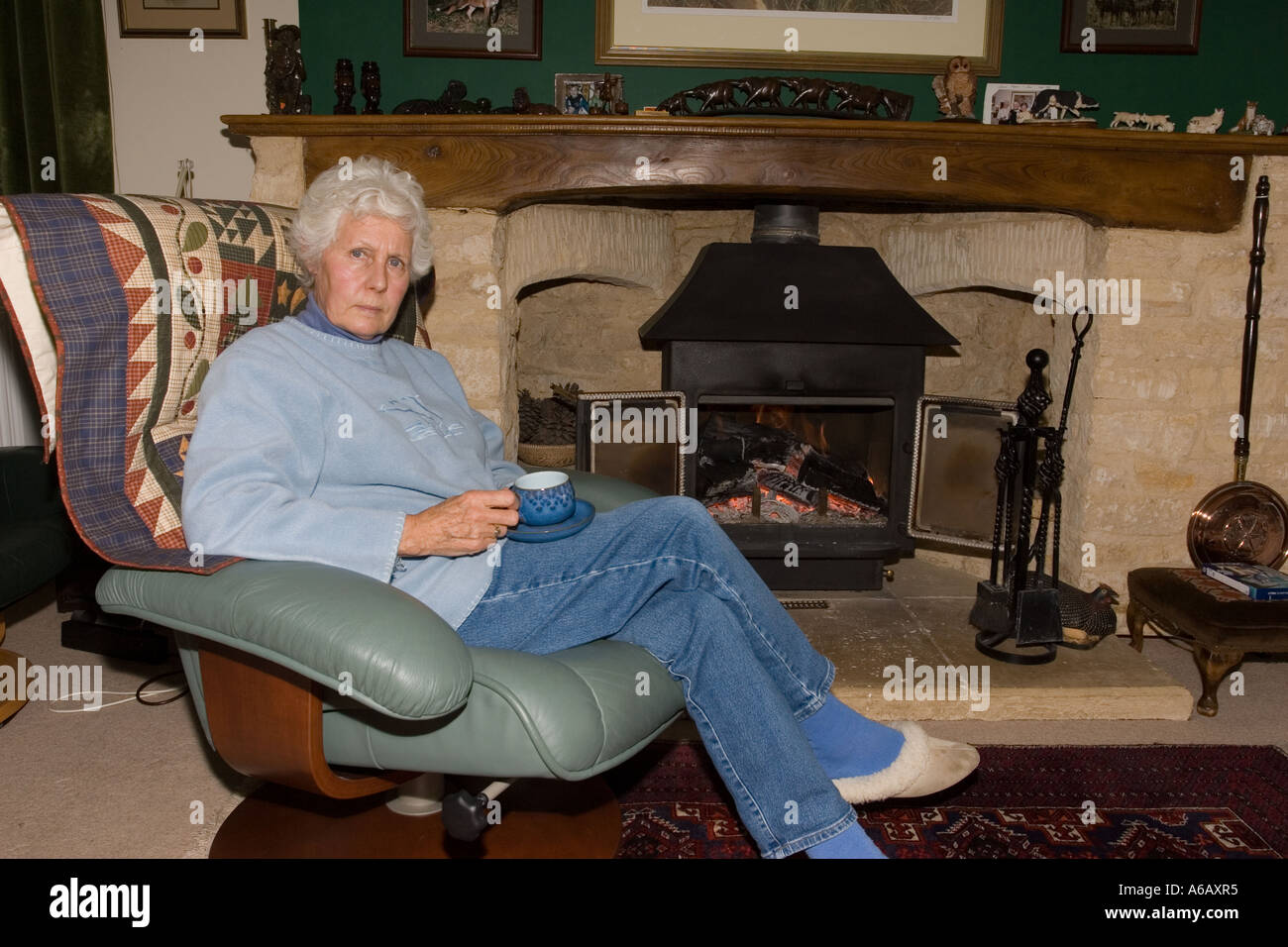 Retired woman relaxing in front of wood fire Cotswolds UK Stock Photo