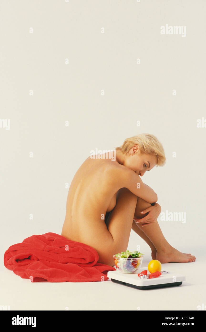 Blond nude woman with short hair seated on red bath robe next to scale and healthy items Stock Photo