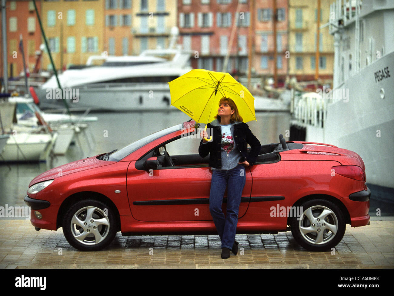 A FEMALE WITH A YELLOW UMBRELLA AND A PEUGEOT 206 COUPE CONVERTIBLE IN ST TROPEZ HARBOUR Stock Photo