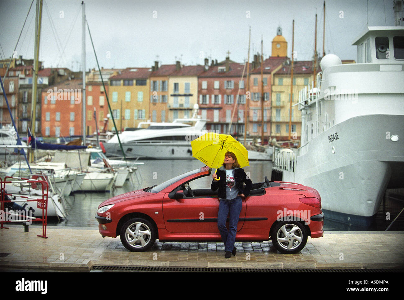 A FEMALE WITH A YELLOW UMBRELLA AND A PEUGEOT 206 COUPE CONVERTIBLE IN ST TROPEZ HARBOUR Stock Photo