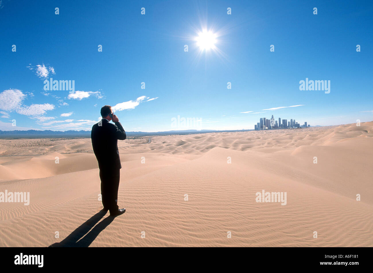 Businessman standing in desolated desert sand dunes talking on cel phone city skyscrapers in the distance  Stock Photo