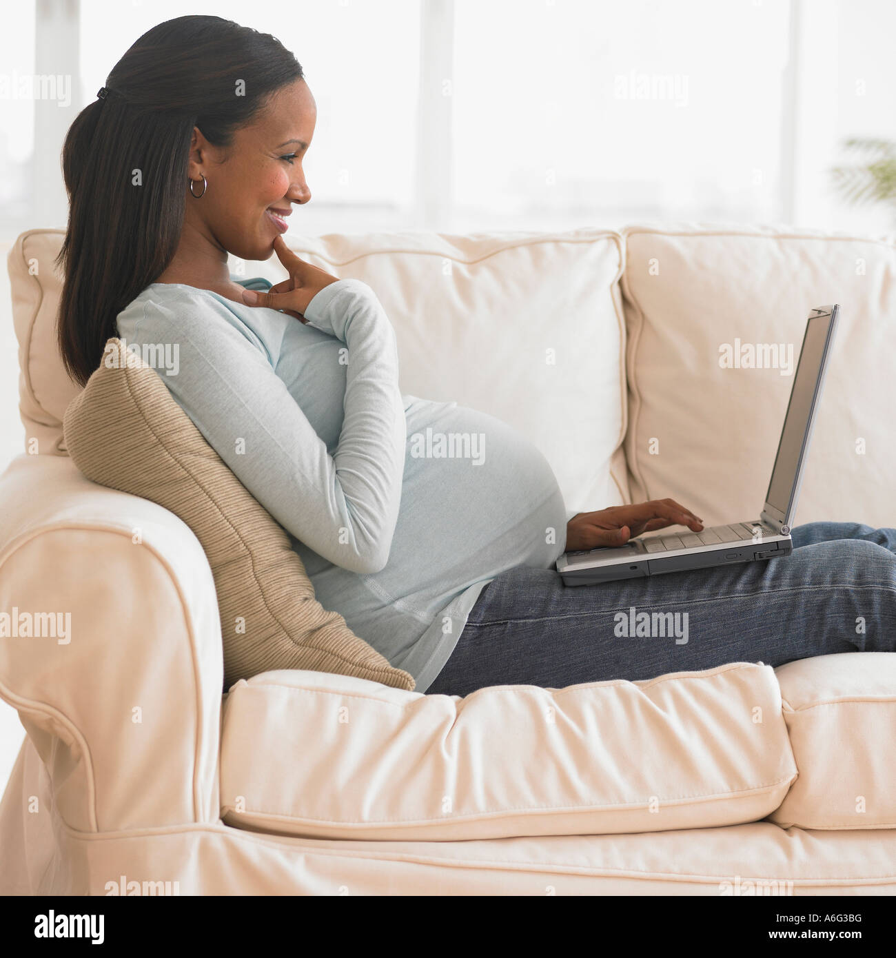 Pregnant African woman typing on laptop on sofa Stock Photo