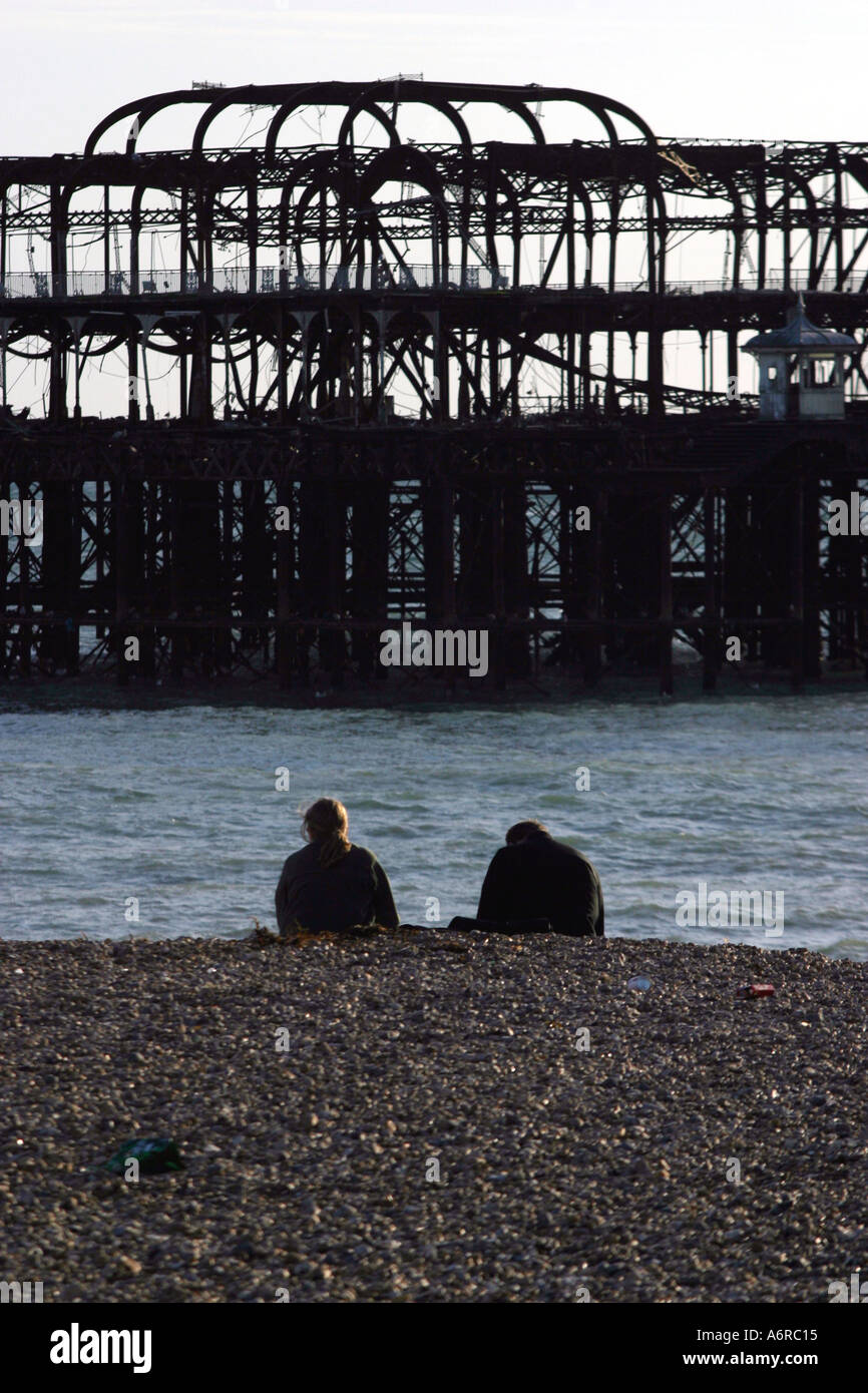 A couple sit on the beach in front of the ruins of Brighton West Pier East Sussex England which was destroyed by fire in 2003 Stock Photo