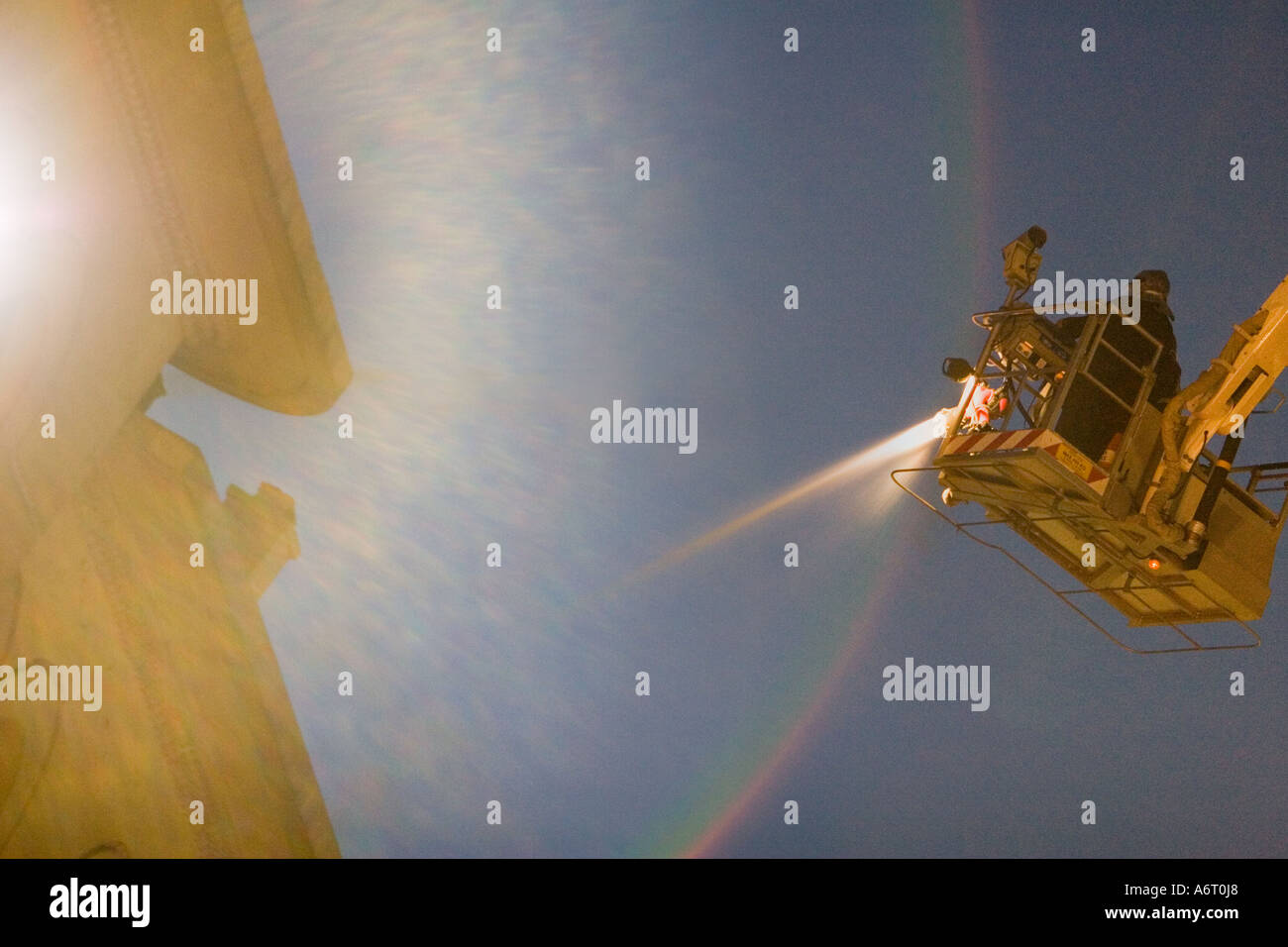 A fireman is throwing water with pressure at fire at a burning building at dusk. The fireman is on a crane. Telephoto view from Stock Photo