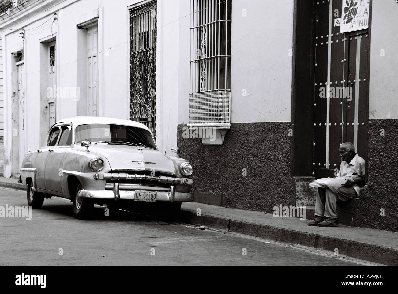 Old man sitting in street near old car in Santiago de Cuba, reading a daily newspaper. Stock Photo