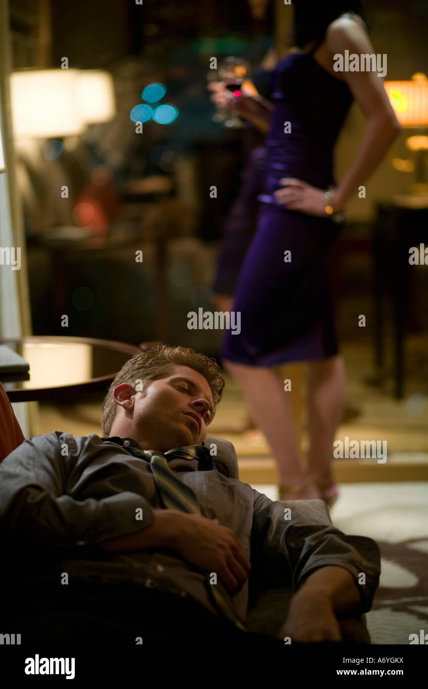 Businessman sleeping on a sofa whilst a woman stands impatiently in the background Stock Photo