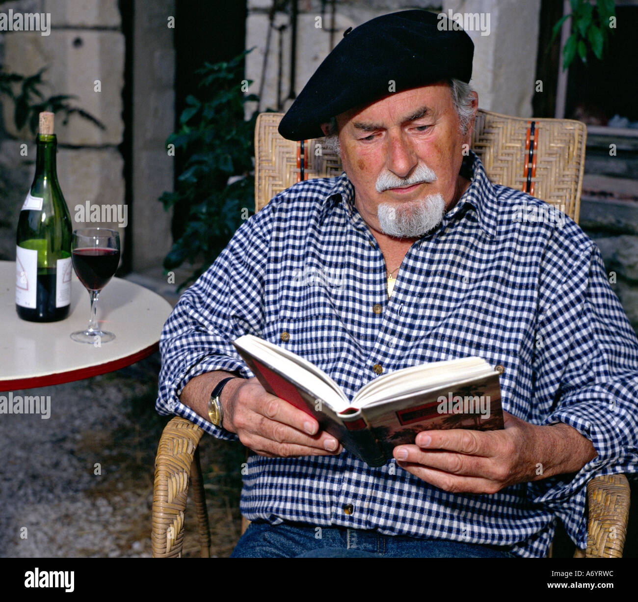 old man reading a book and sitting on a terrace of an old stone house in southern France Stock Photo