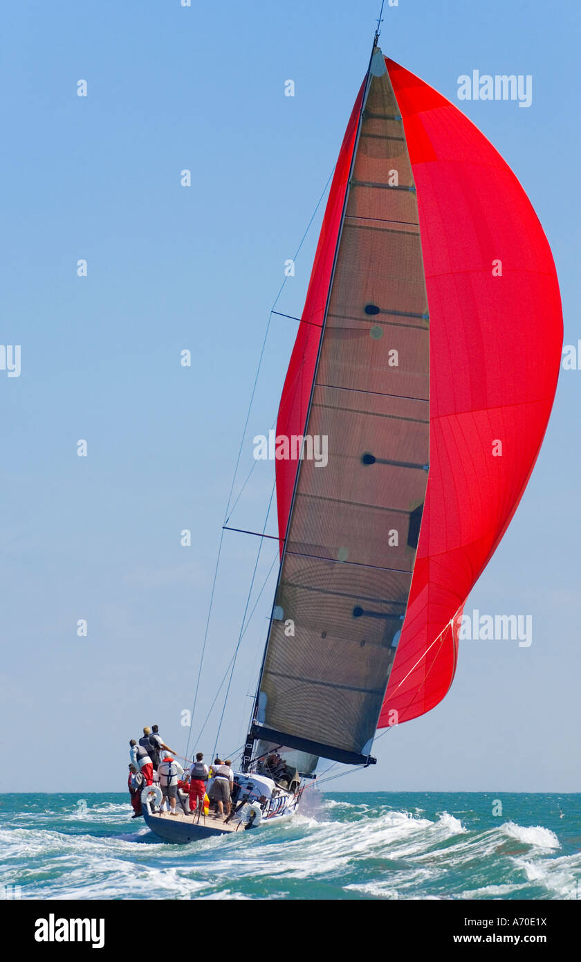 A fully crewed racing yacht with a red spinnaker catching the wind and leaving a big wake Stock Photo