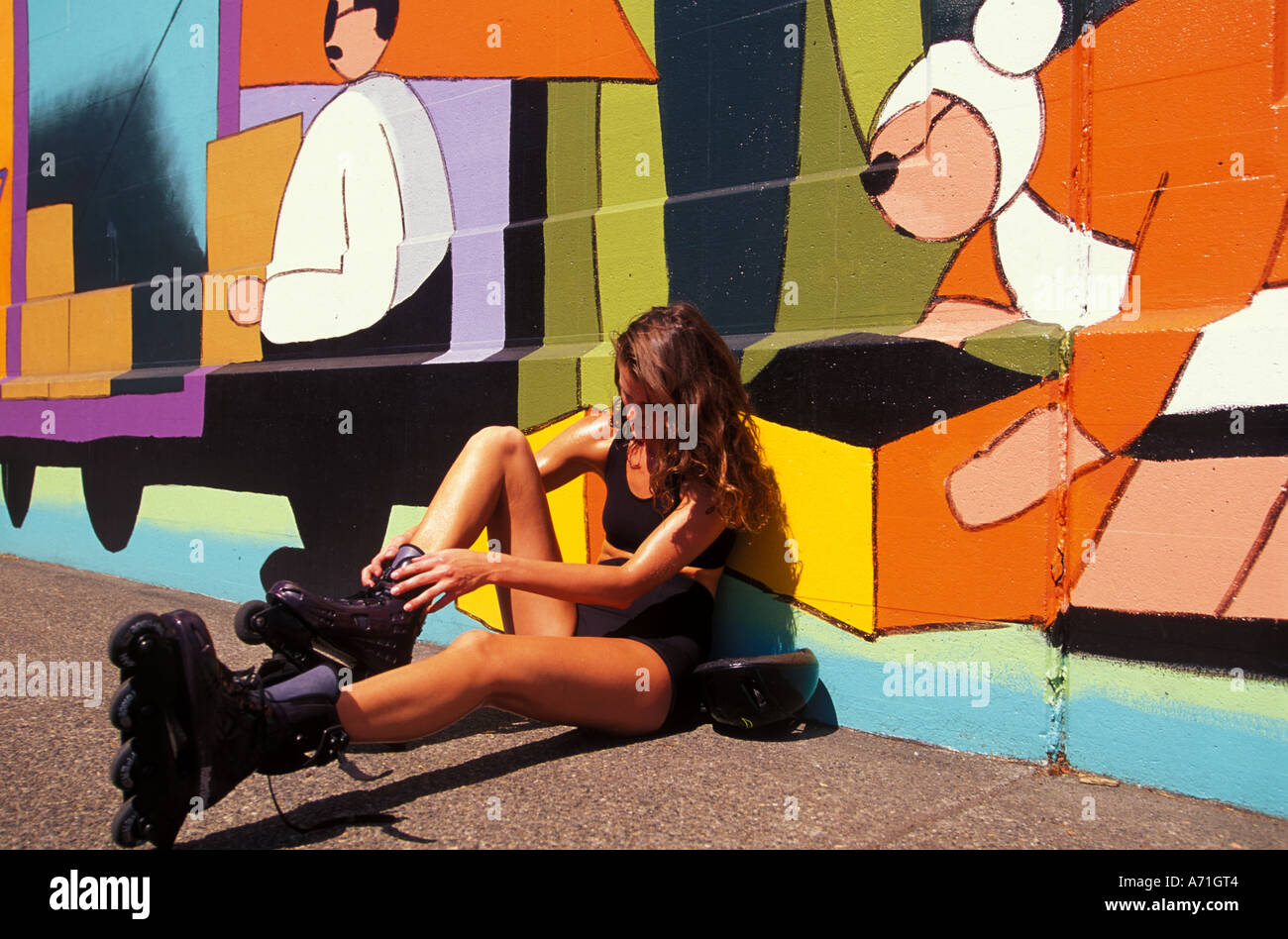 A young woman laces up her in line skates as she sits against against a colorfully painted mural Stock Photo