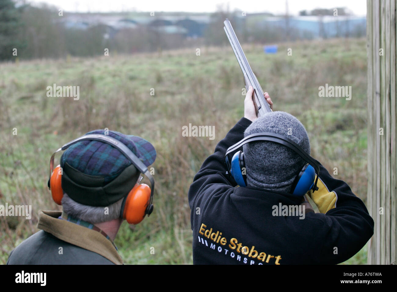 man wearing blue ear defenders in shooting area takes aim with shotgun under instruction on