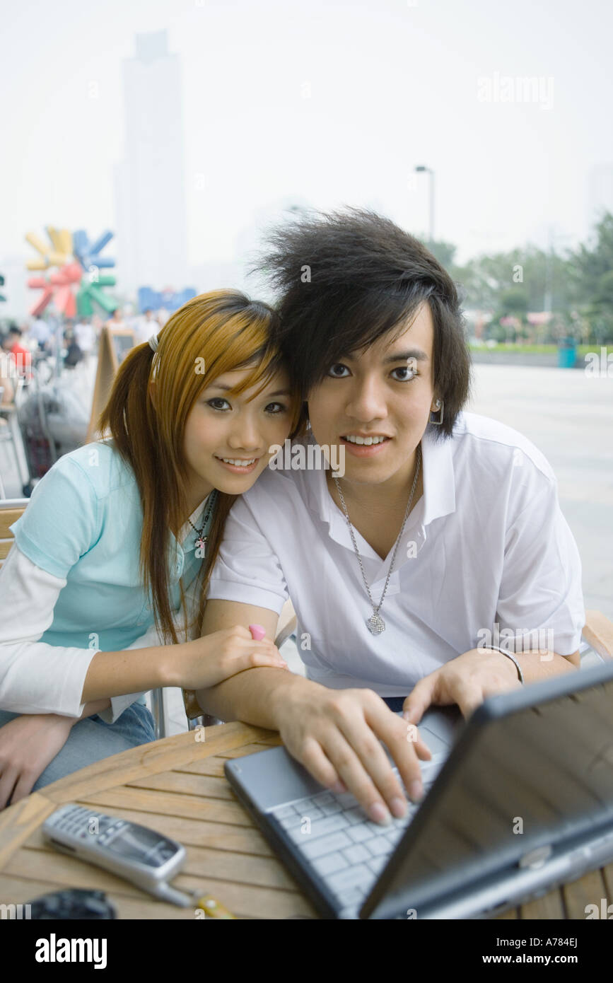 Teenage couple sitting outdoors, using laptop, smiling at camera Stock Photo