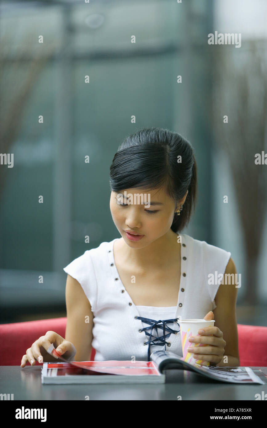 Young woman sitting at table, looking at magazine, holding drink Stock Photo