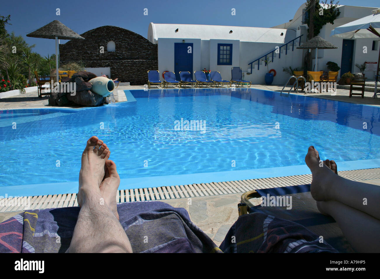 The legs of two people, a couple resting on beach chairs near a blue swimming pool Acrotiri Santorini Greece Stock Photo