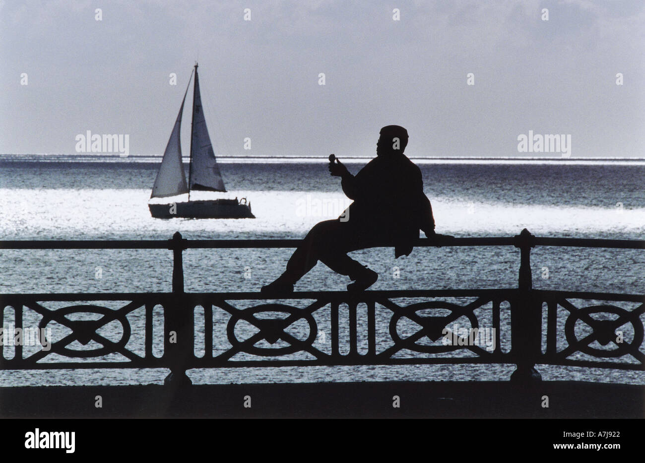 Seafront of the city of Brighton and Hove. A plump woman sits on railings eating ice cream as a yacht  sails past. Stock Photo