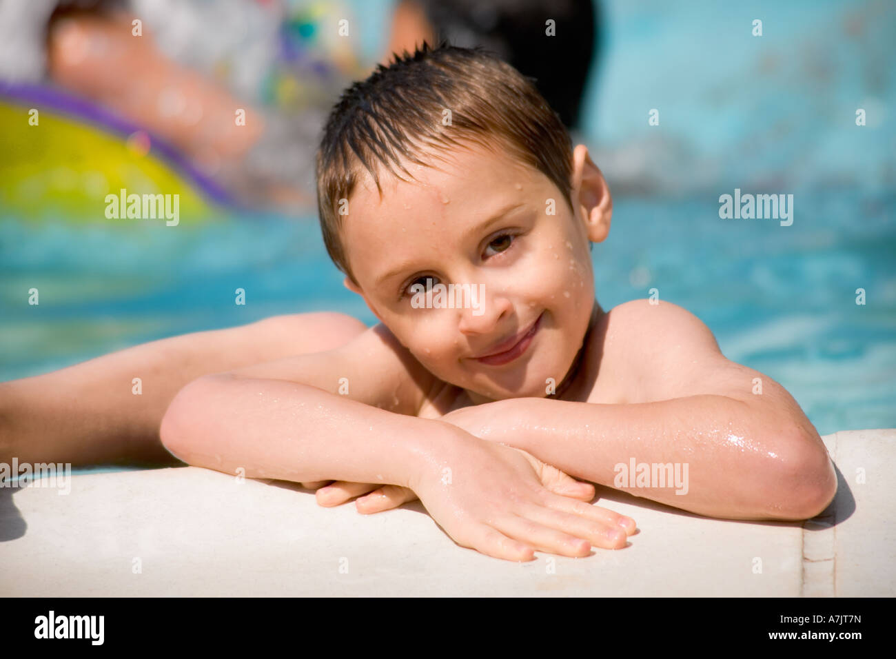 wet Caucasian boy in a swimming pool resting at poolside smiling at the camera with beads of water on his face Stock Photo