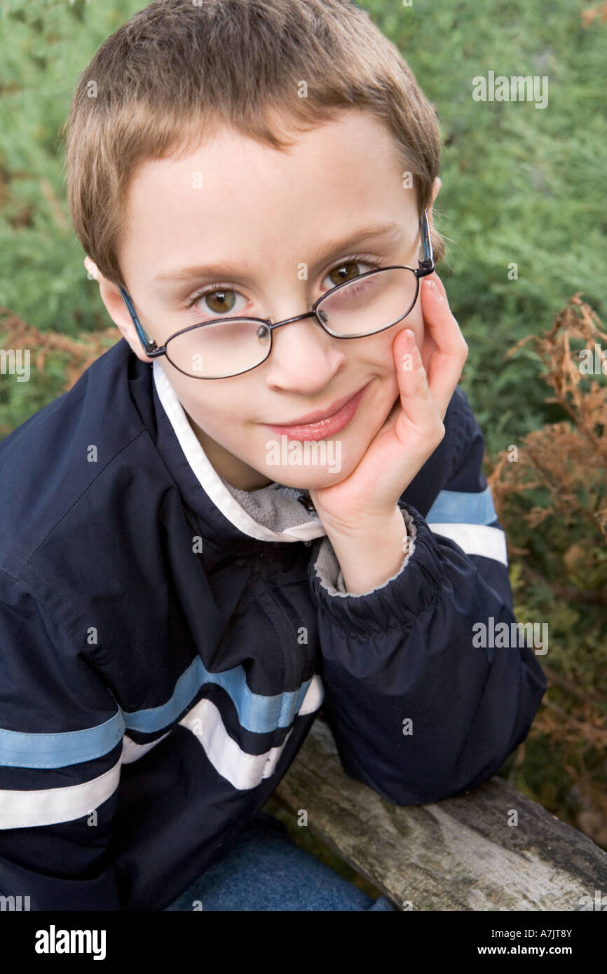 portrait of young boy wearing glasses and light jacket outdoors leaning his chin in his open hand elbow on a fence rail Stock Photo