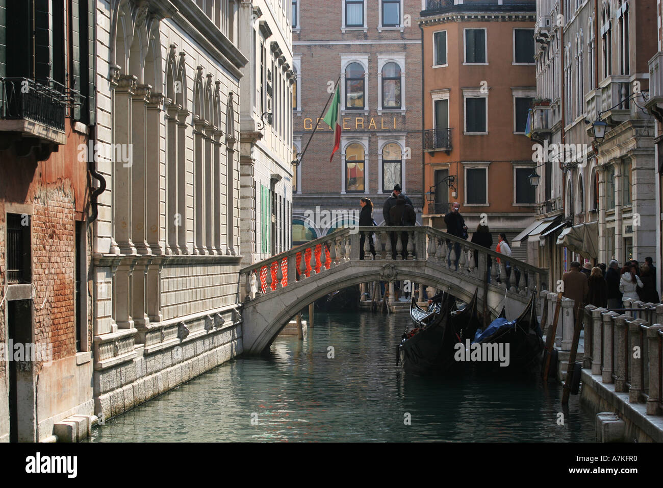 Tourists crowd a small backstreet bridge over a tyical Venetian canal with Italian flag and famous gondolar boats Venice Italy Stock Photo