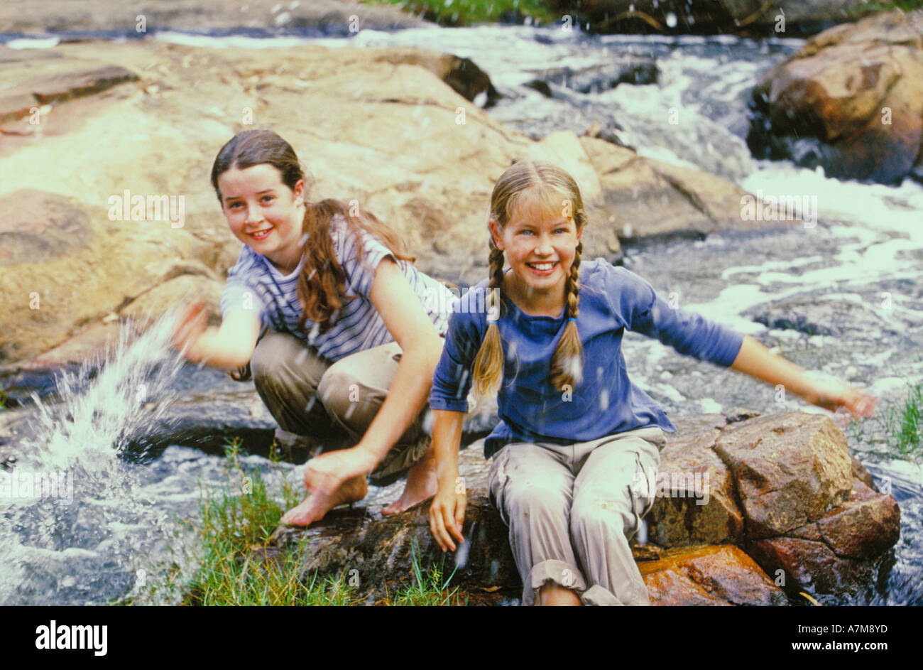 two female teenager sitting on a stone rock in a river sputtering water ...