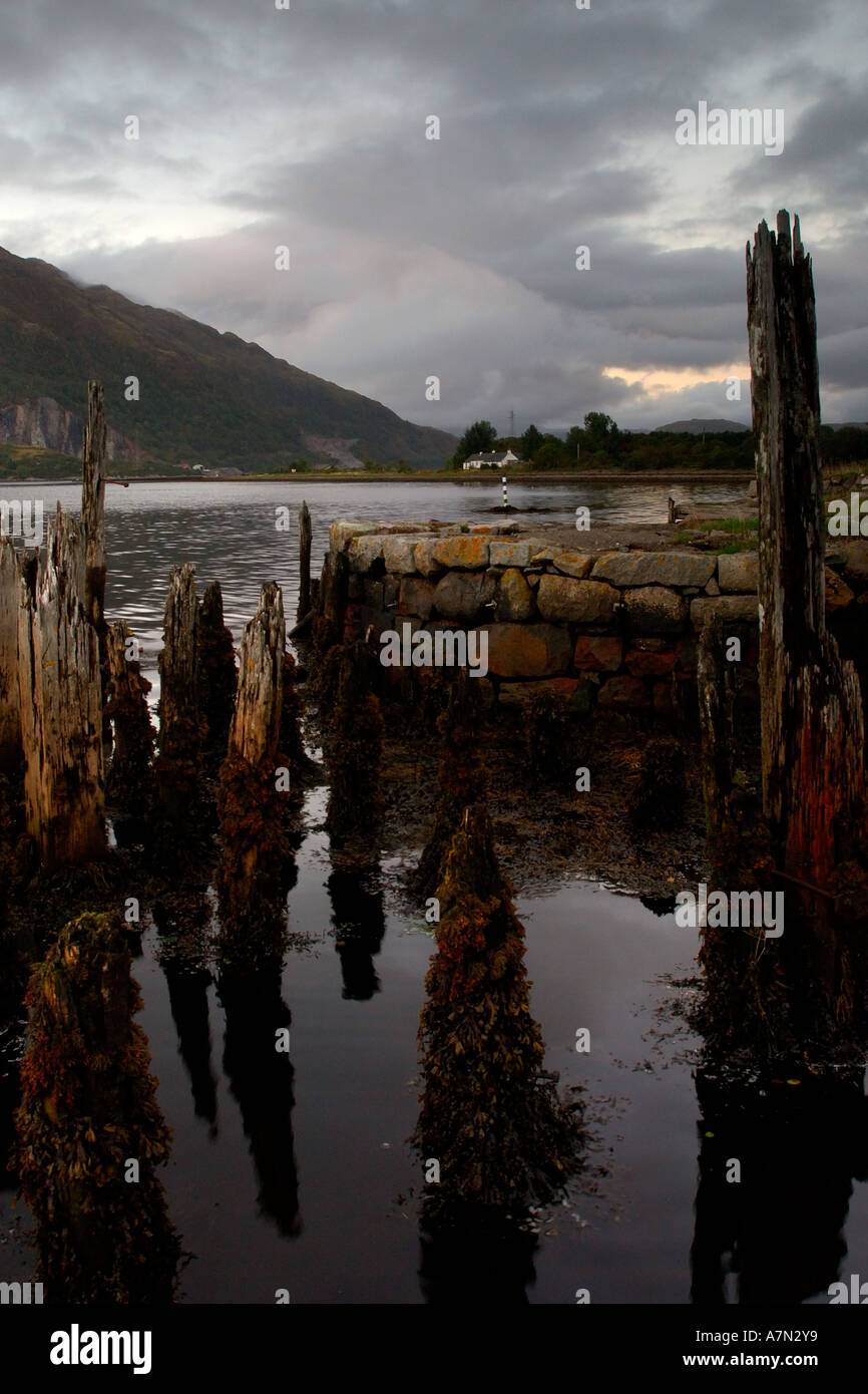Rotting wooden jetty posts at the end of a stone built victorian pier with overcast threatening stormy sky Loch Etive Scotland Stock Photo