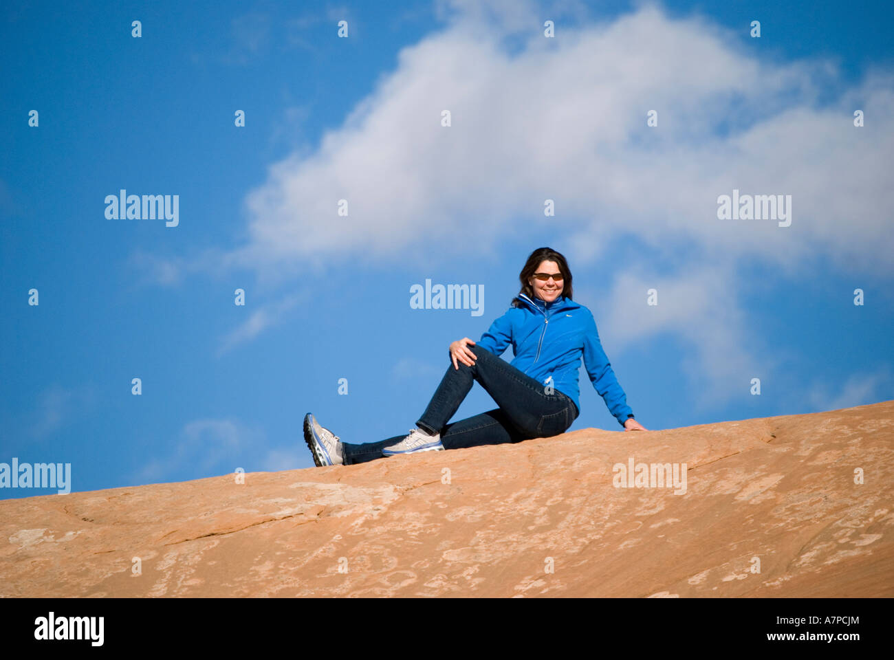 Smiling woman hiker sitting on Entrada sandstone ridge with blue sky and white clouds Arches National Park Utah USA Stock Photo
