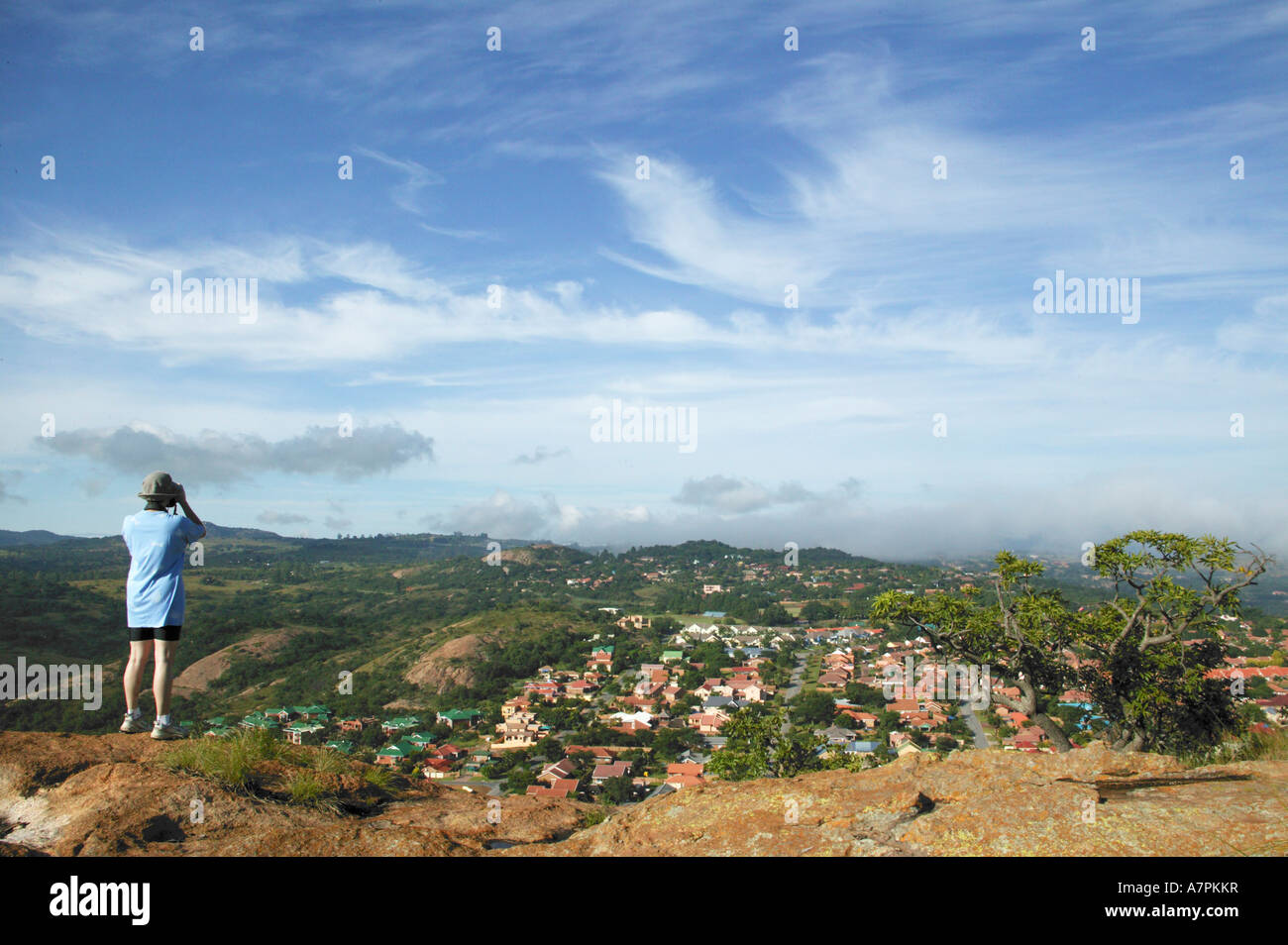 A person standing on the edge of a granite outcrop looking through binoculars towards a residential suburb in the valley below Stock Photo