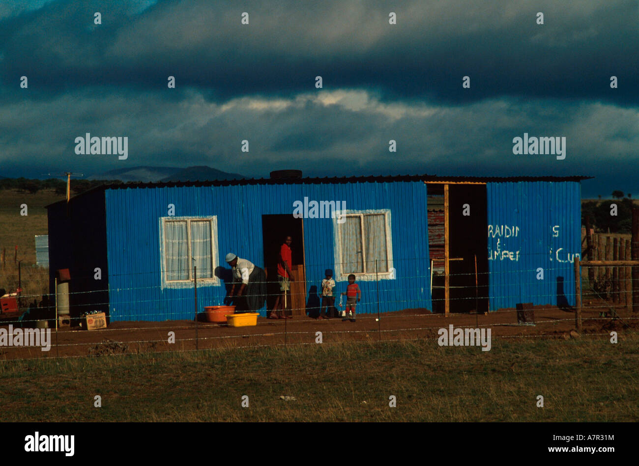 A family works outside a blue corrugated iron shack dwelling against a stormy sky with residents in the doorway Stock Photo