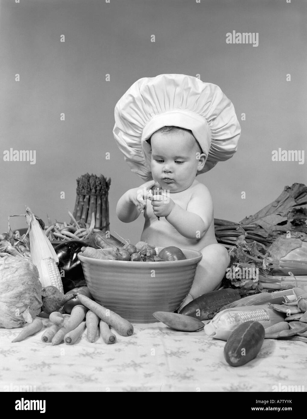 1950s 1960s BABY WEARING CHEF HAT TOQUE BOWL AND RAW VEGETABLES MAKING A SALAD Stock Photo