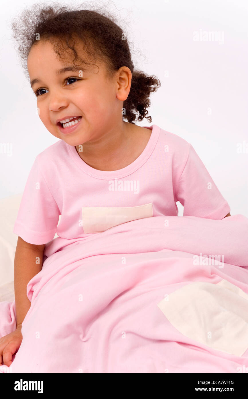 A beautiful little mixed race girl dressed in pink pajamas sitting up in bed and smiling Stock Photo
