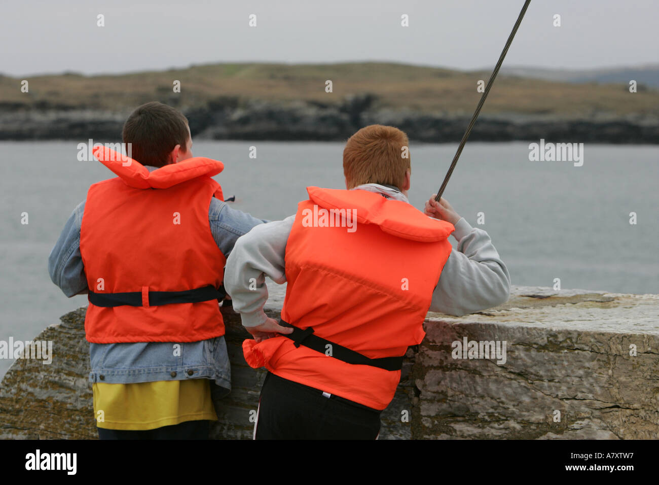 two young teenage boys wearing orange hi vis life jackets waiting for a boat to come in on Downings pier Stock Photo