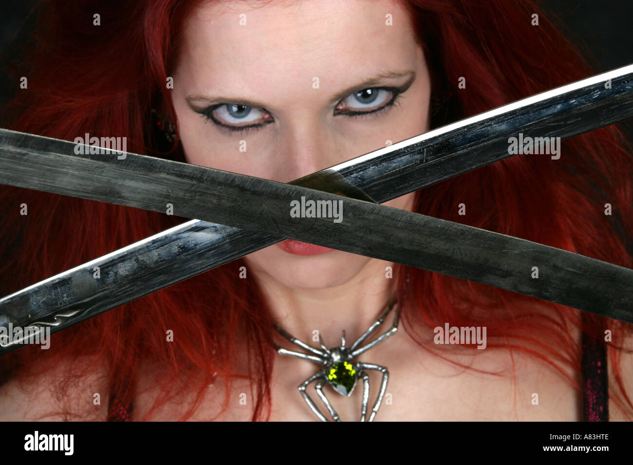 Woman with Red Hair Wearing a Spider Necklace and Holding Two Crossed Swords in Front of Her Face Stock Photo