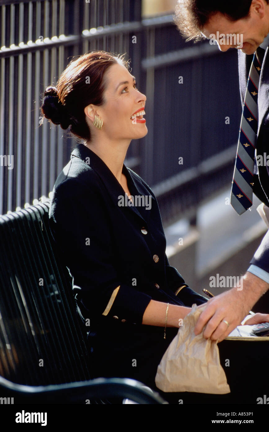 Close-up of smartly dressed woman sitting on bench smiling up at man in suit standing outdoors in urban setting. Stock Photo