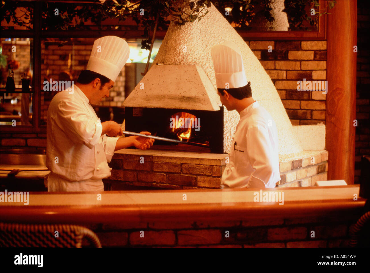 Two men hotel chefs in whites tending pizza oven in restaurant. Stock Photo