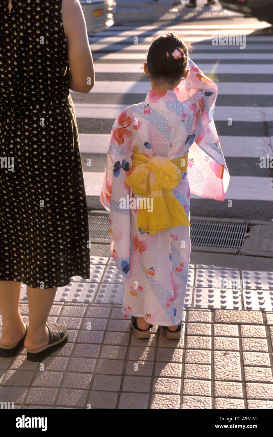 A little girl dressed in a cute summer yukata robe with yellow sash waiting with her mother to cross the street Stock Photo