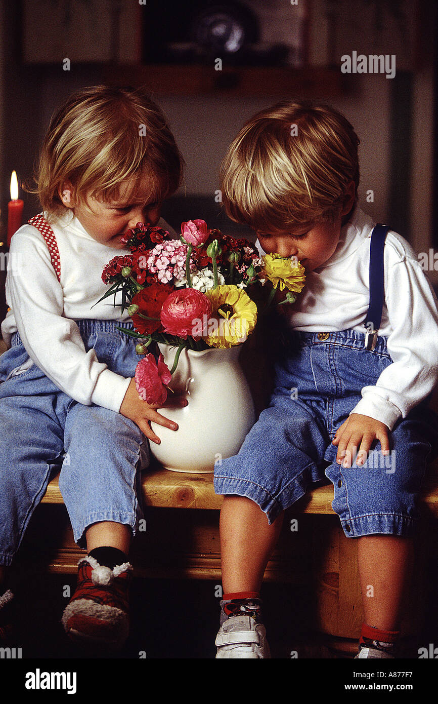Two young children sitting on a bench in jeans suspenders and white shirts smelling a base of fresh flowers Stock Photo