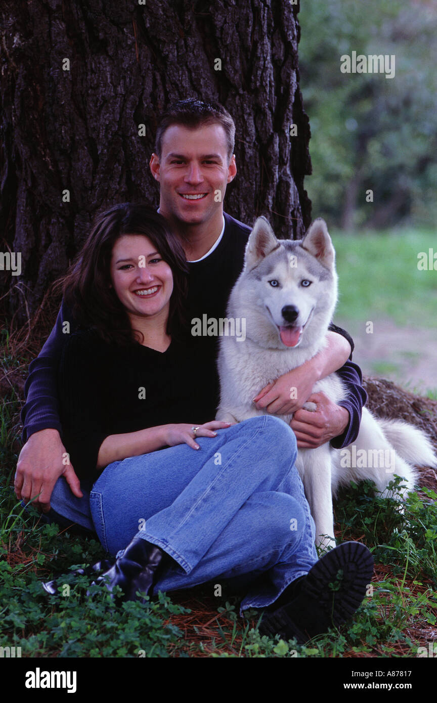 A young couple sitting under a tall tree with their husky dog Stock Photo