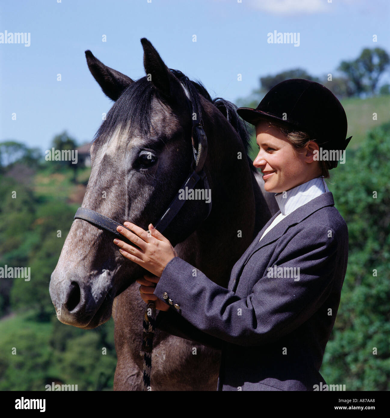 A smiling young woman in a formal gray riders outfit and black helmet strokes the face of her horse as she holds its bridle Stock Photo