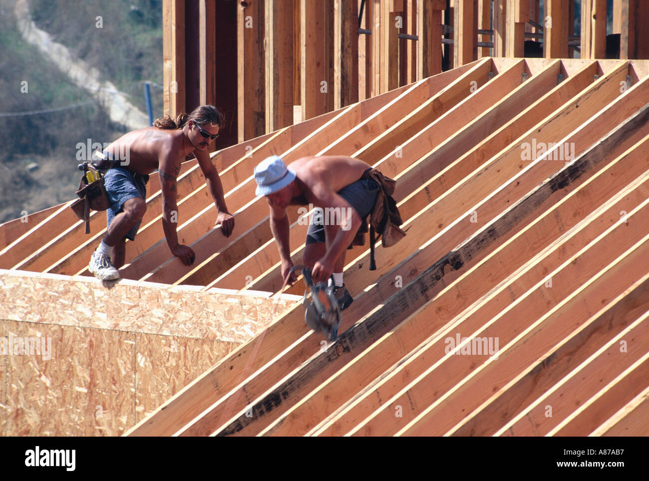 Two shirtless carpenters working on the roof framing of a large wooden