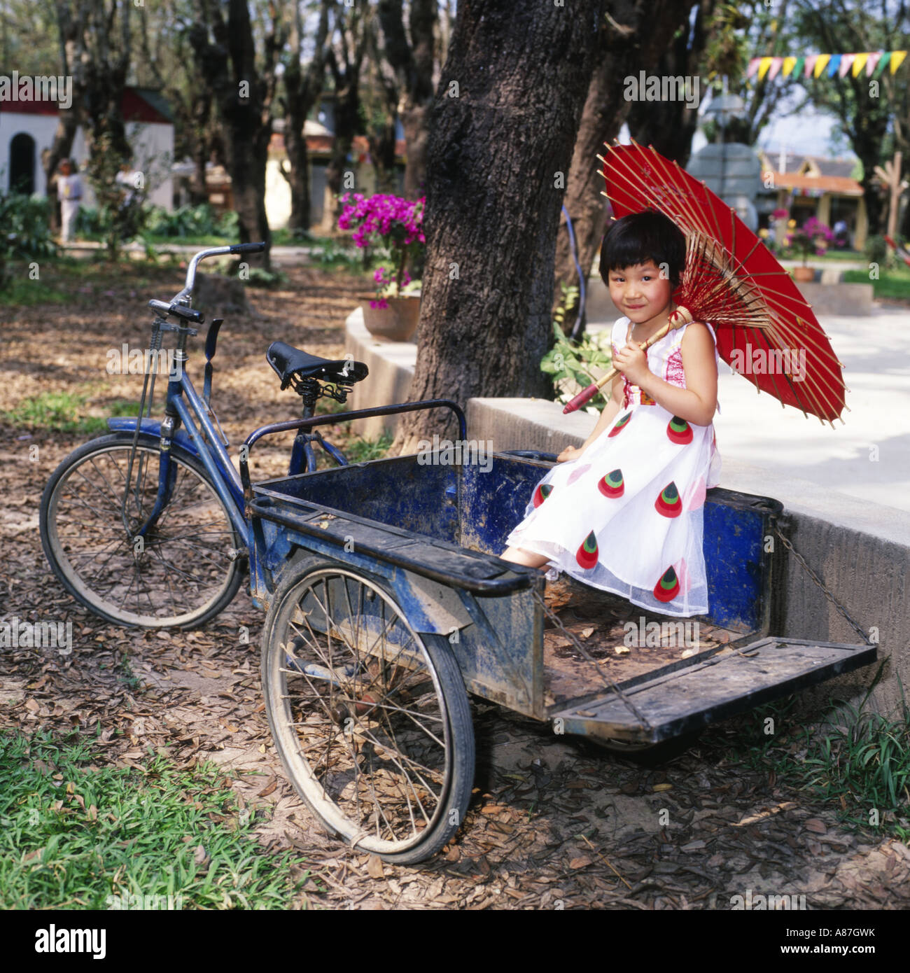 Young local girl,holding red parasol,sitting in the back of tricycle trailer,Jinghong,Yunnan province,China Stock Photo