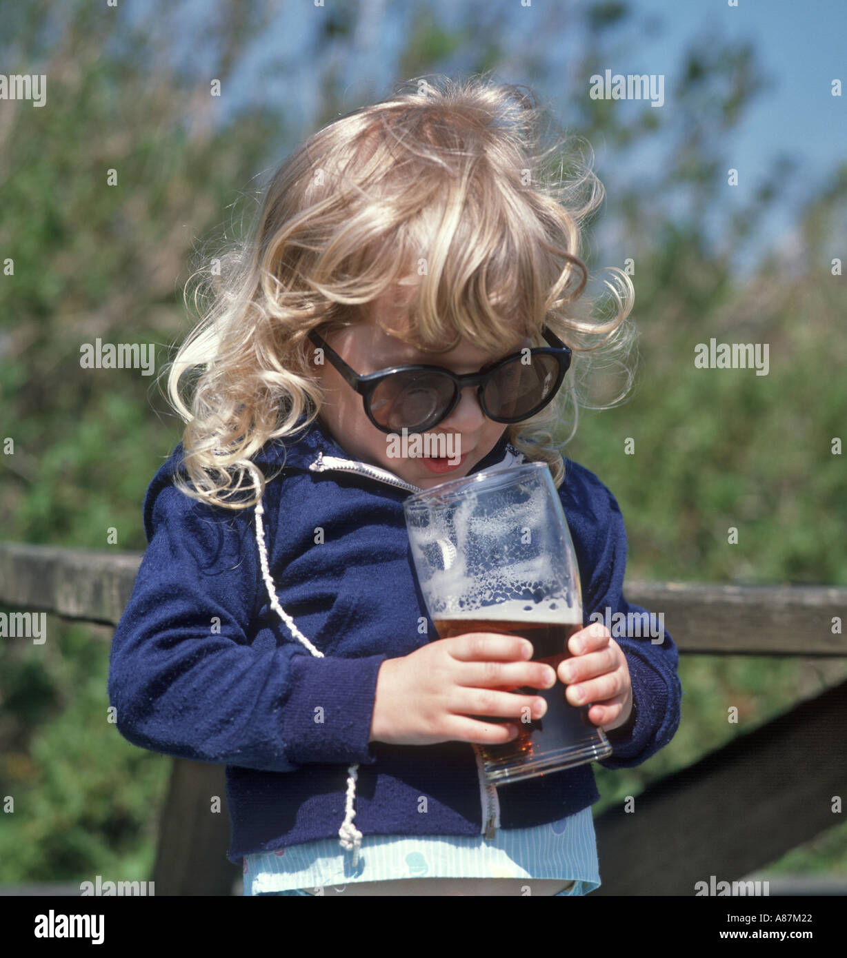 Little Girl trying to drink a pint of beer and wearing oversized sunglasses, England, United Kingdom Stock Photo