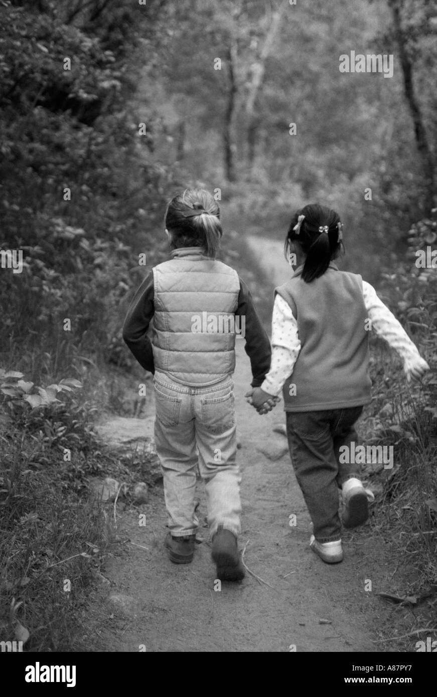 Black and white image of two young girls holding hands and hiking on a path through the forest Stock Photo