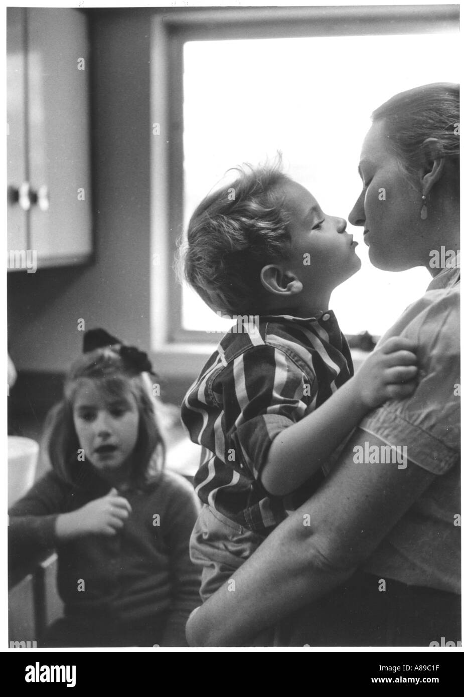 Younger brother gives mother a kiss while older sister looks on Stock Photo