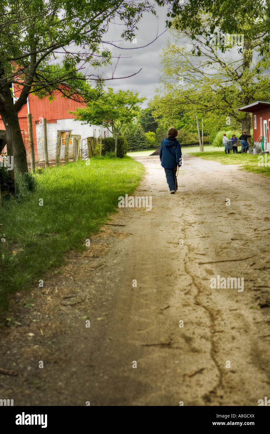 boy on farm walking down dirt lane dragging a stick that makes a line along his pathway Stock Photo