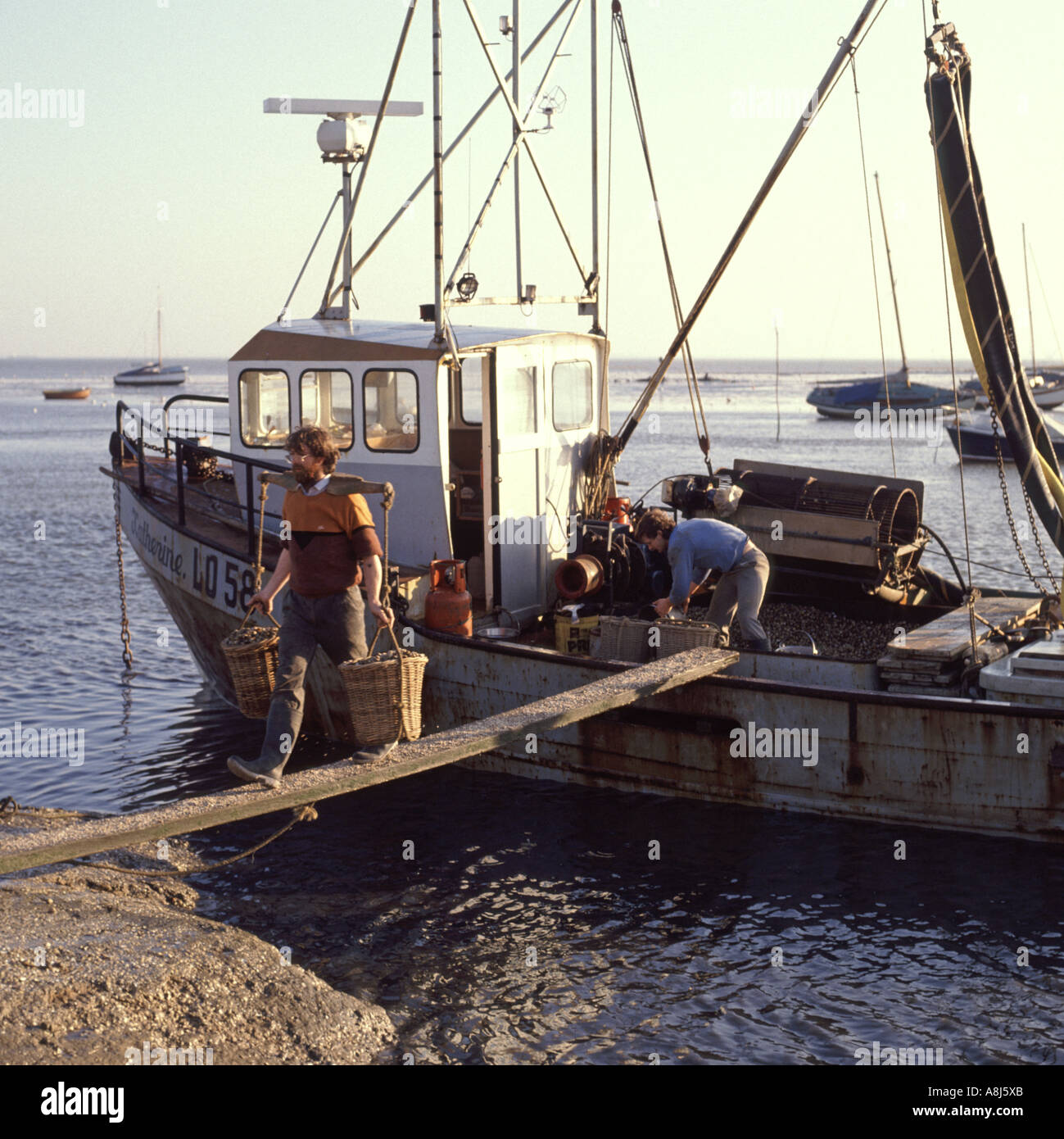 1980s archive view men at work on plank using shoulder yokes to carry shell fish baskets from fishing boat 80s Leigh on Sea Southend Essex England UK Stock Photo