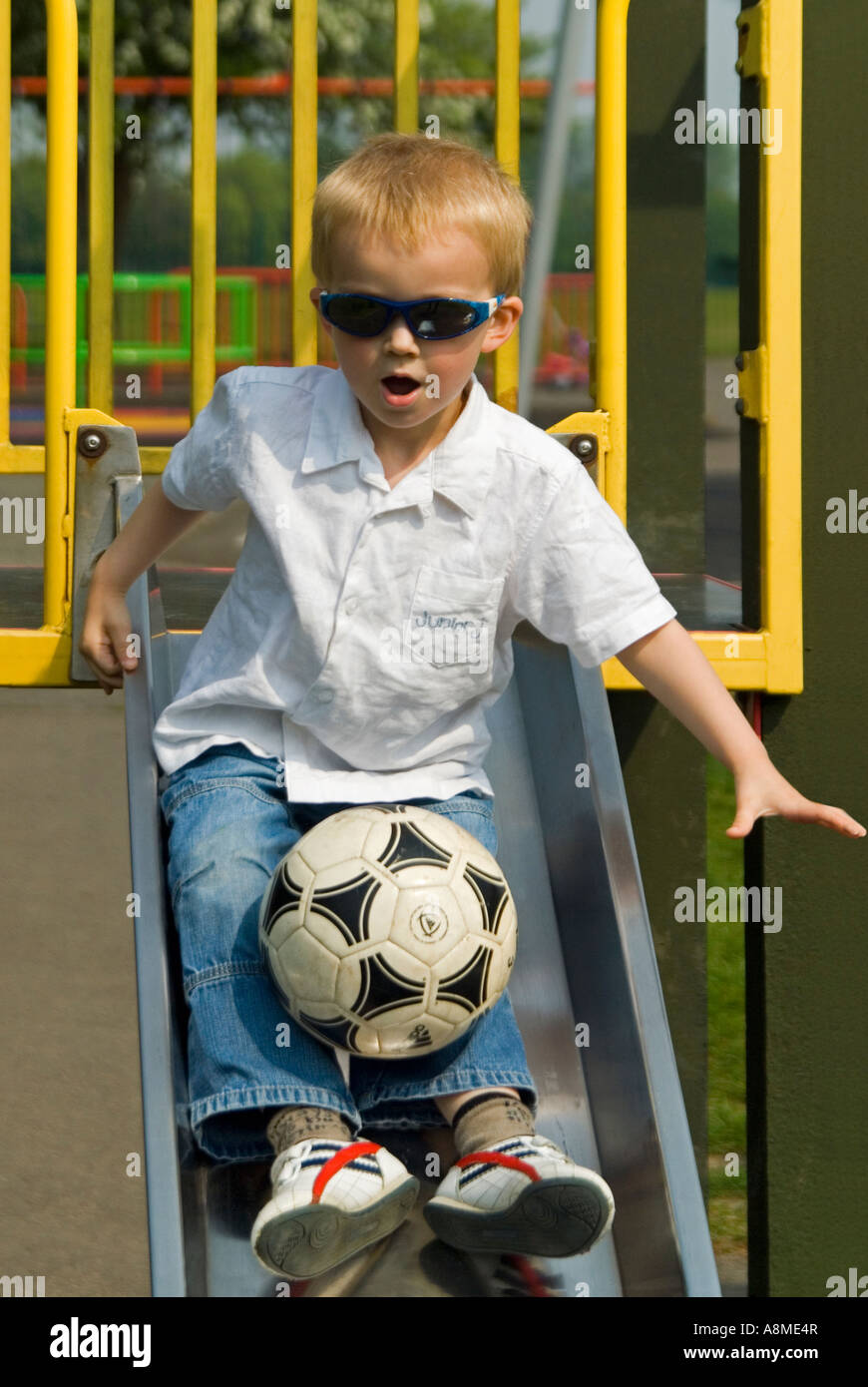 Vertical portrait of a young boy (4 year old) at the top of a slide, sliding down feet first with a football on his lap. Stock Photo