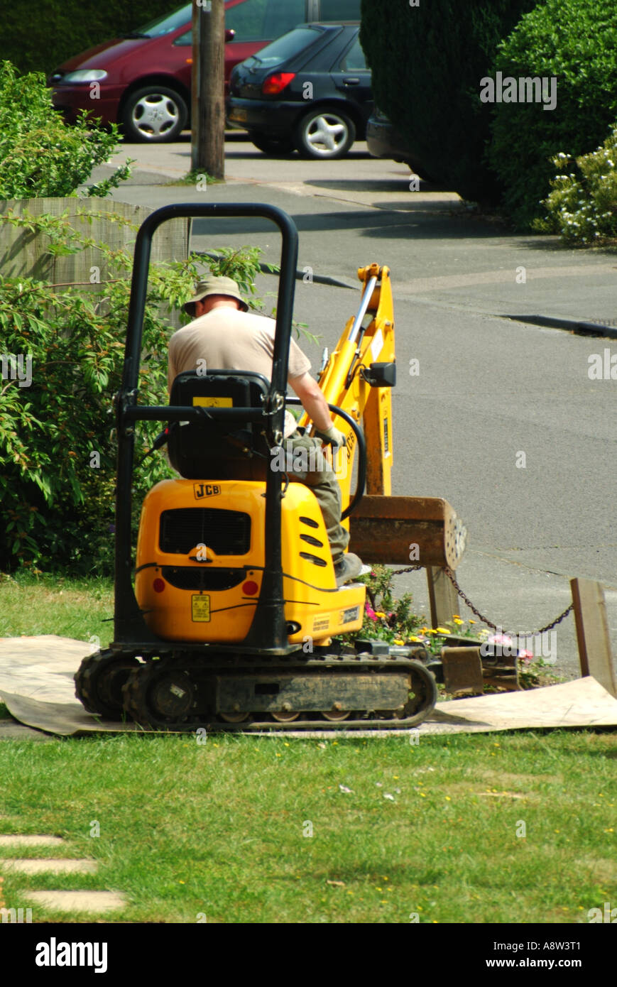 Man driving mini track laying excavator crawling across domestic front garden lawn over plywood protection sheets Essex England UK Stock Photo