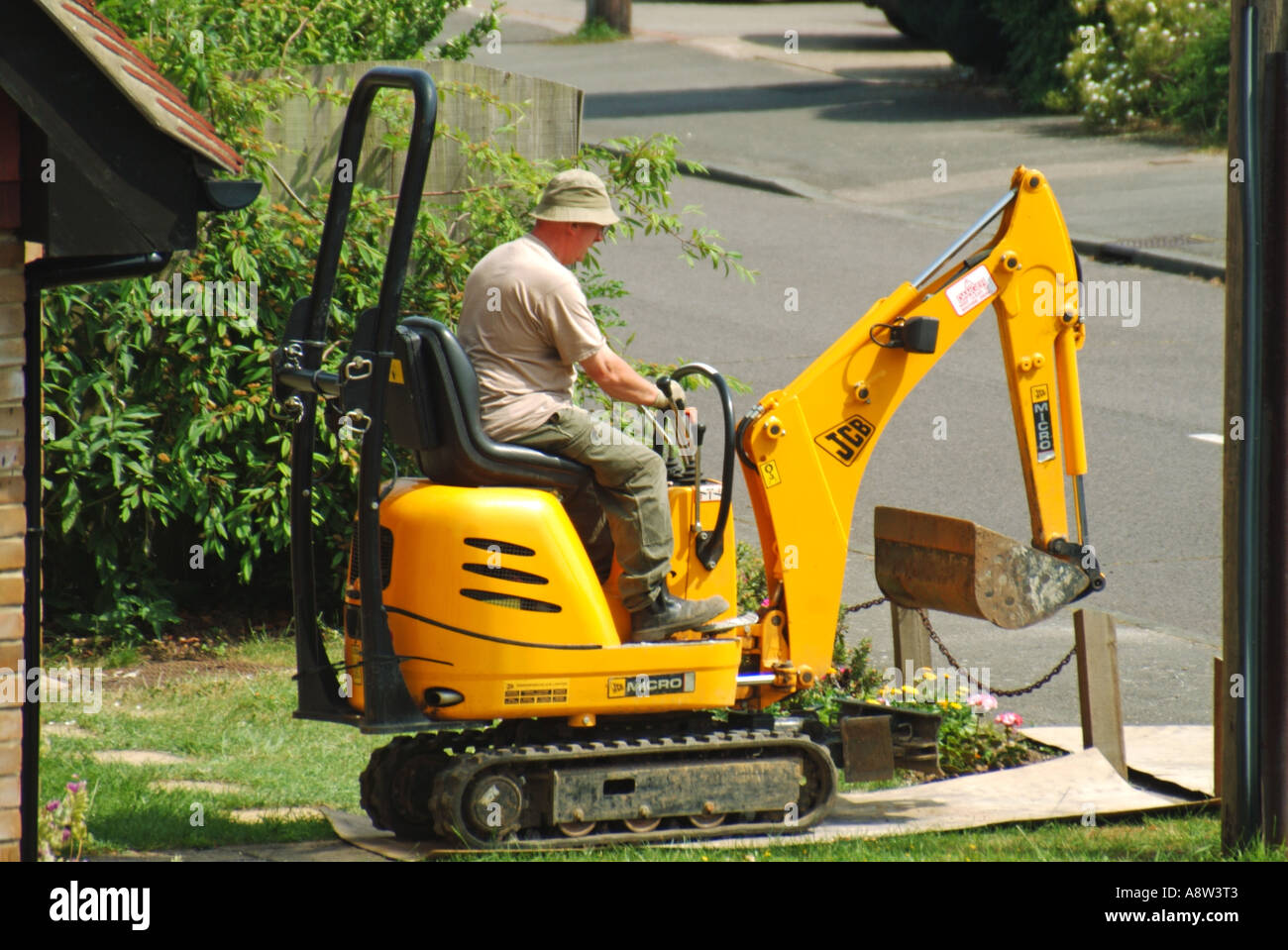 Man driving mini track laying excavator crawling across domestic front garden lawn over plywood protection sheets Essex England UK Stock Photo