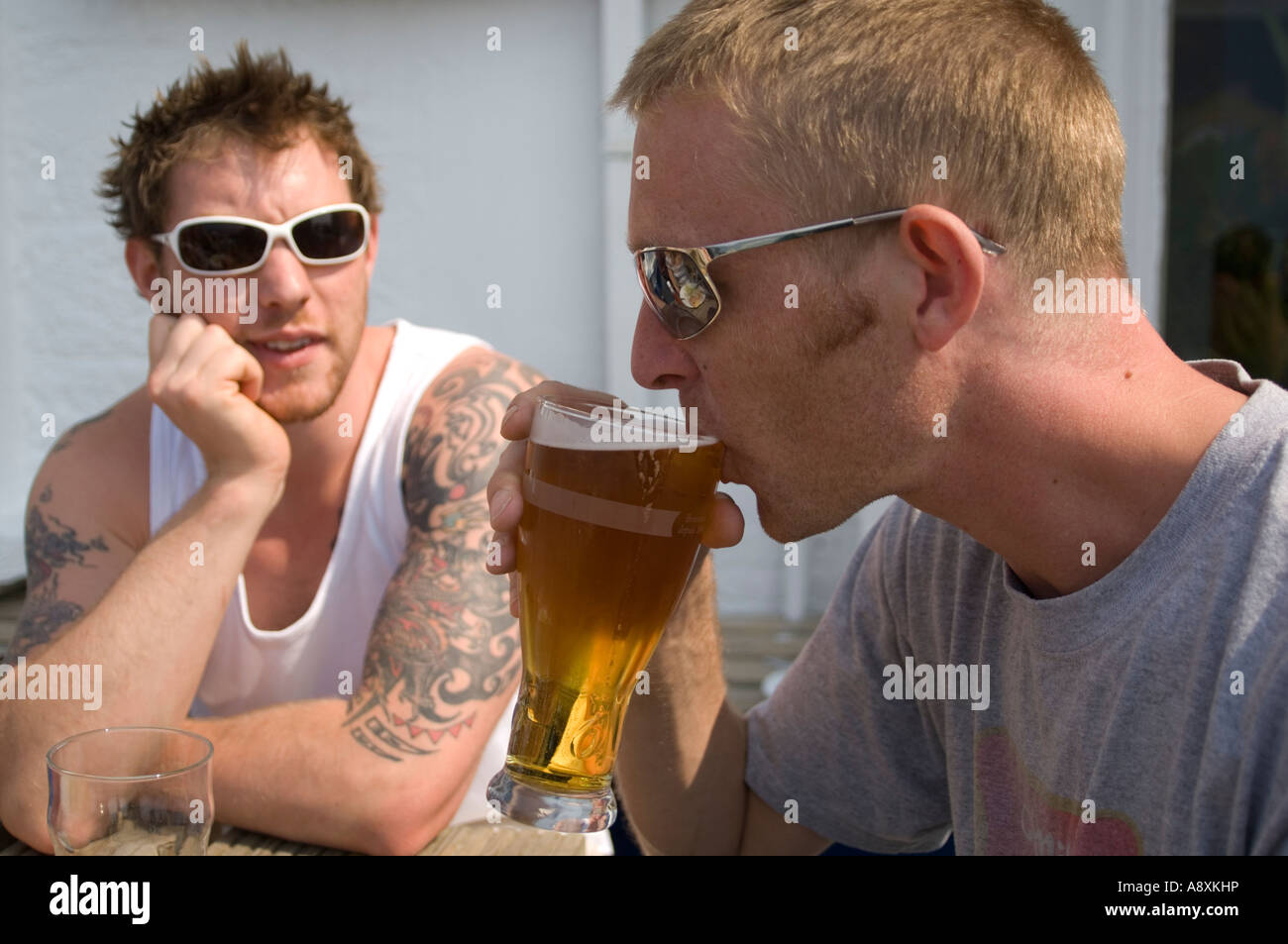 Man drinking pint of beer at outside table of pub as friend with heavy tribal tattoos looks at him, UK Stock Photo